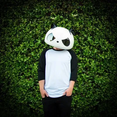 A person wearing a large, stylized panda mask with black and white features stands in front of a green leafy hedge.