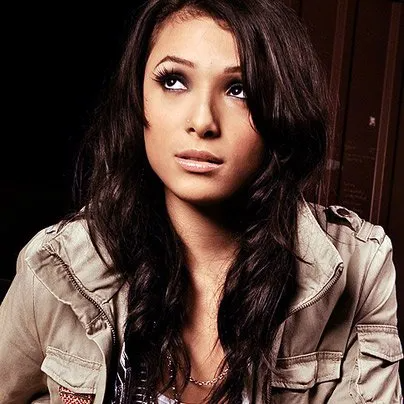 A young person with long dark wavy hair, wearing a tan jacket, looking upward with a thoughtful expression.