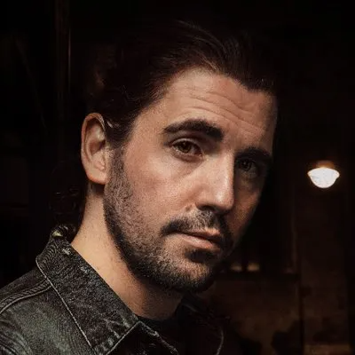 A person with dark hair and a beard, wearing a denim jacket, looks directly at the camera in a dimly lit setting.