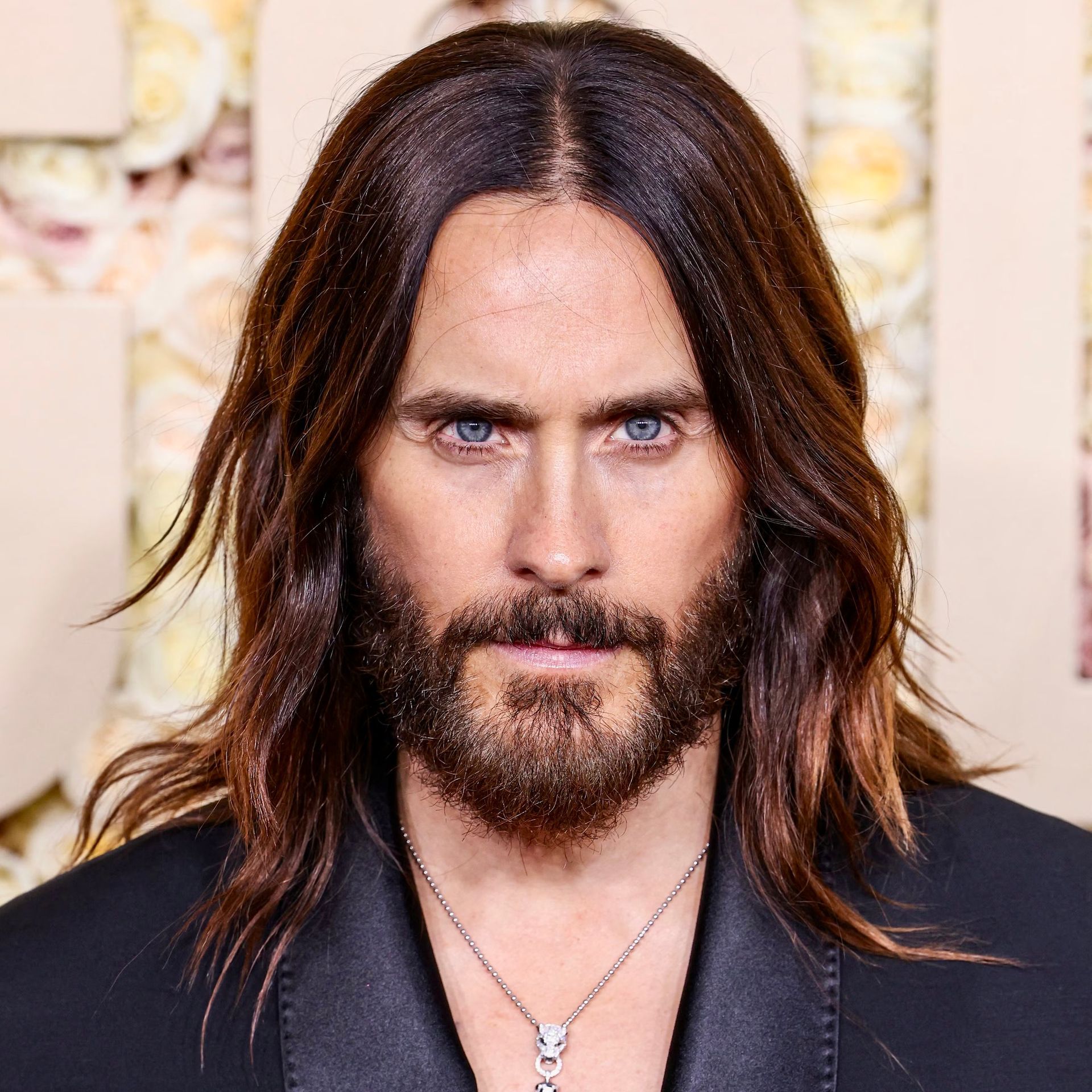 A close-up portrait of Jared Leto with shoulder-length hair and a beard, wearing a black blazer and a silver pendant.