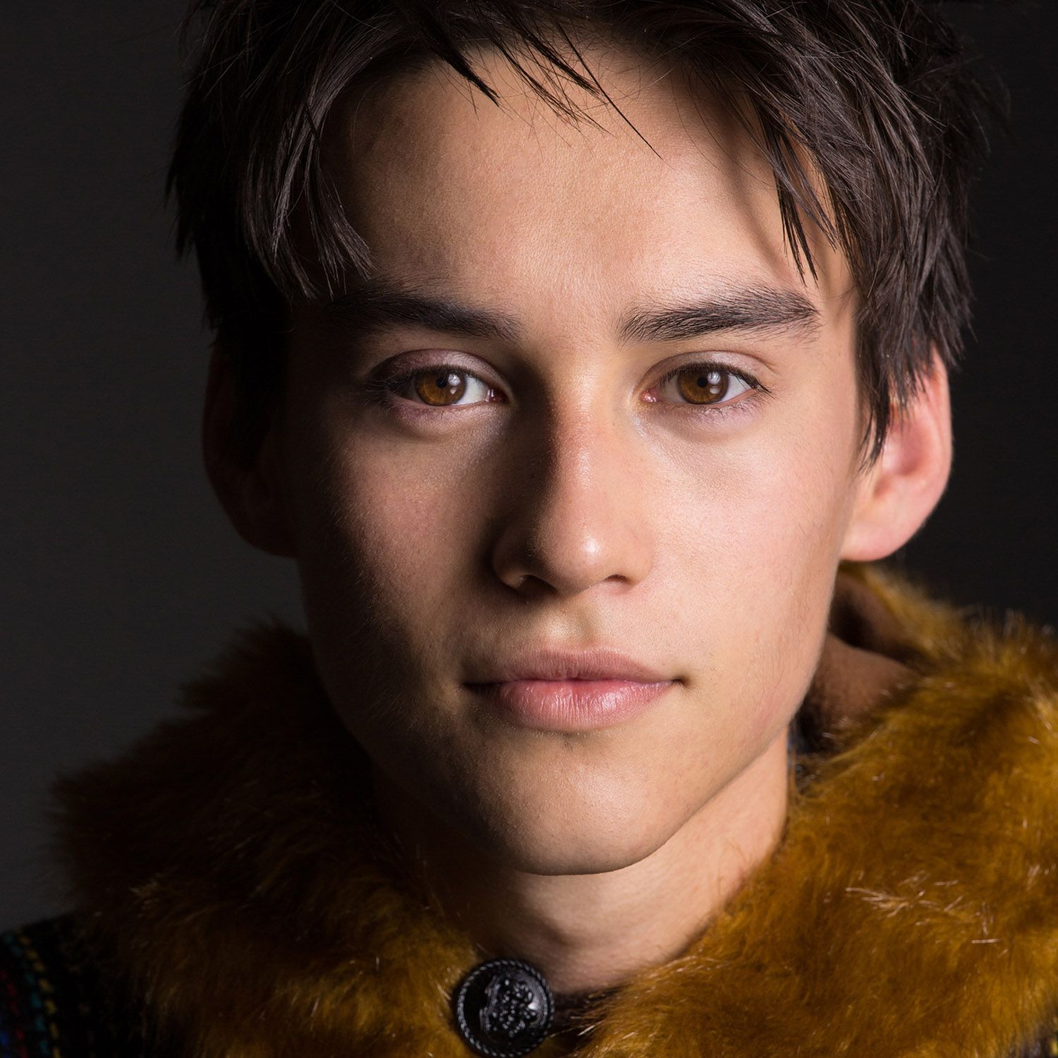A close-up portrait of a person with brown eyes and dark hair, wearing a dark garment with a mustard-yellow fur collar.