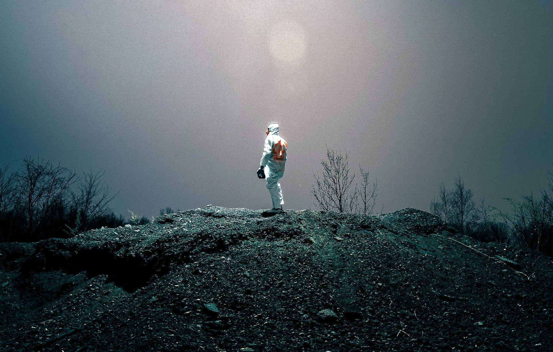 A person in a white protective suit stands on a dark, rocky hill under a bright, misty sky filled with falling particles.