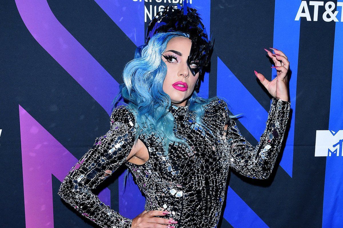 Lady Gaga with wavy blue and black hair, wearing a shimmering silver long-sleeved outfit against a branded blue backdrop.