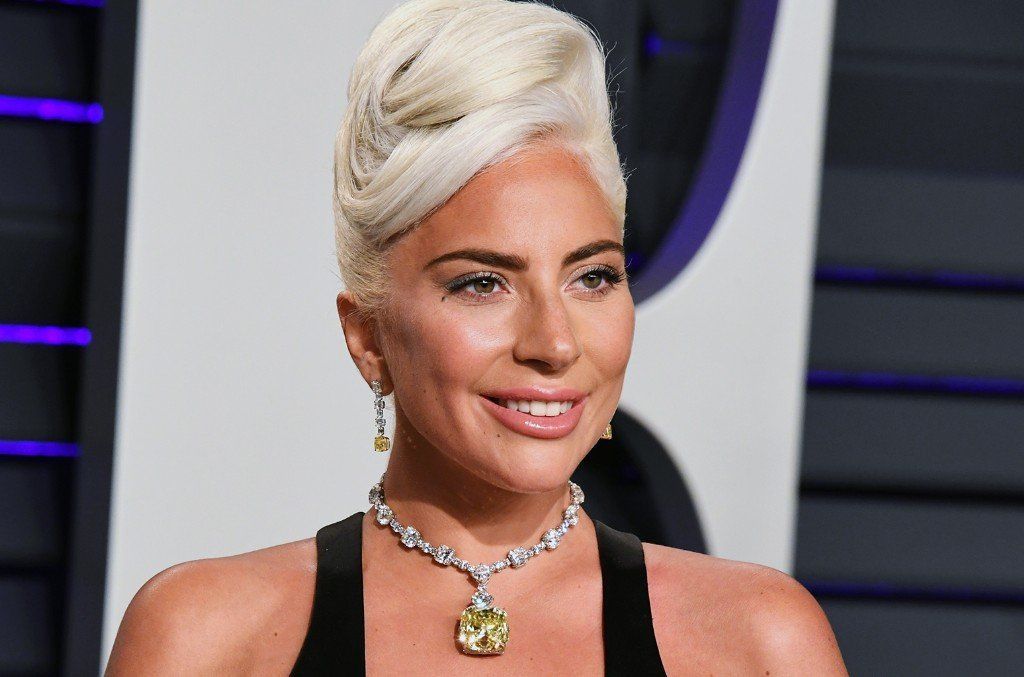 Lady Gaga poses in a black dress with a yellow diamond pendant necklace and an elaborate blonde updo at a formal event.