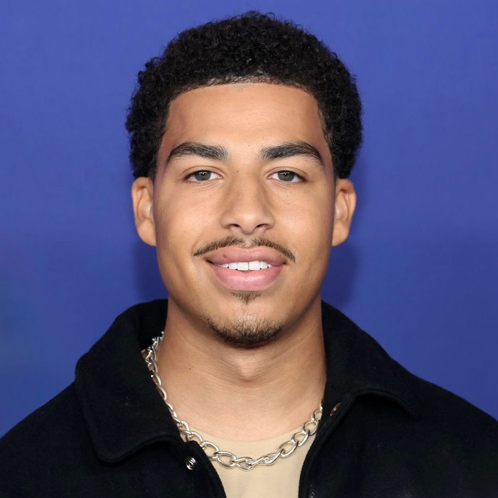 A person with dark curly hair and a mustache smiles against a plain, solid blue background.