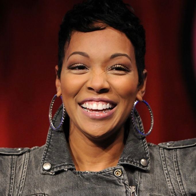 A smiling person with short dark hair, wearing a grey denim jacket and large circular earrings against a red background.