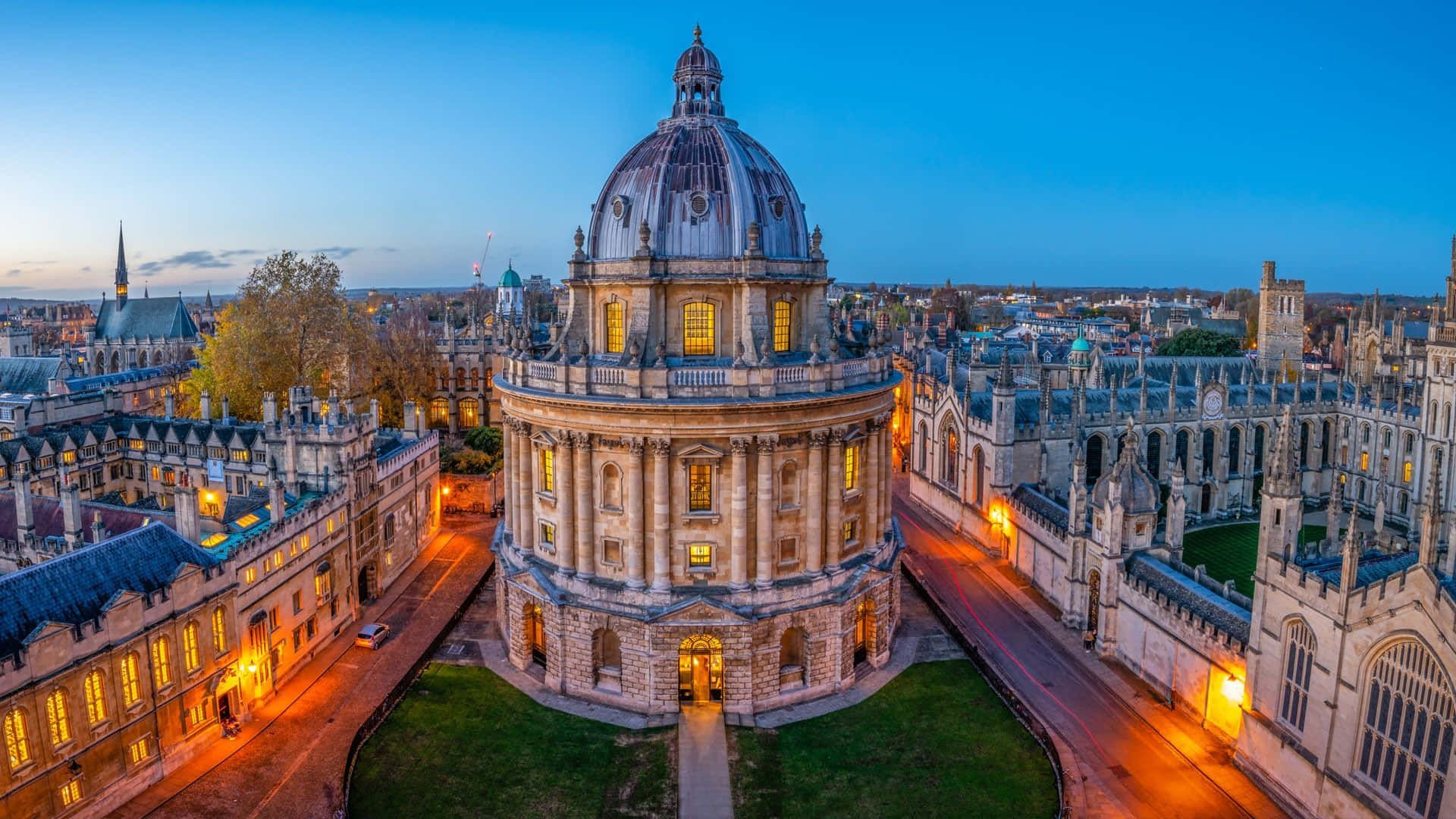 Aerial view of the Radcliffe Camera in Oxford, England, illuminated by warm lights against a blue twilight sky.