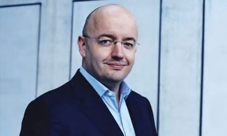 A person with a shaved head and glasses wearing a dark blazer and blue dress shirt, standing before a light blue wall.