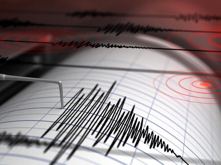A seismograph needle records sharp, erratic black waves on a grid paper roll, with red ripples visible in the background.
