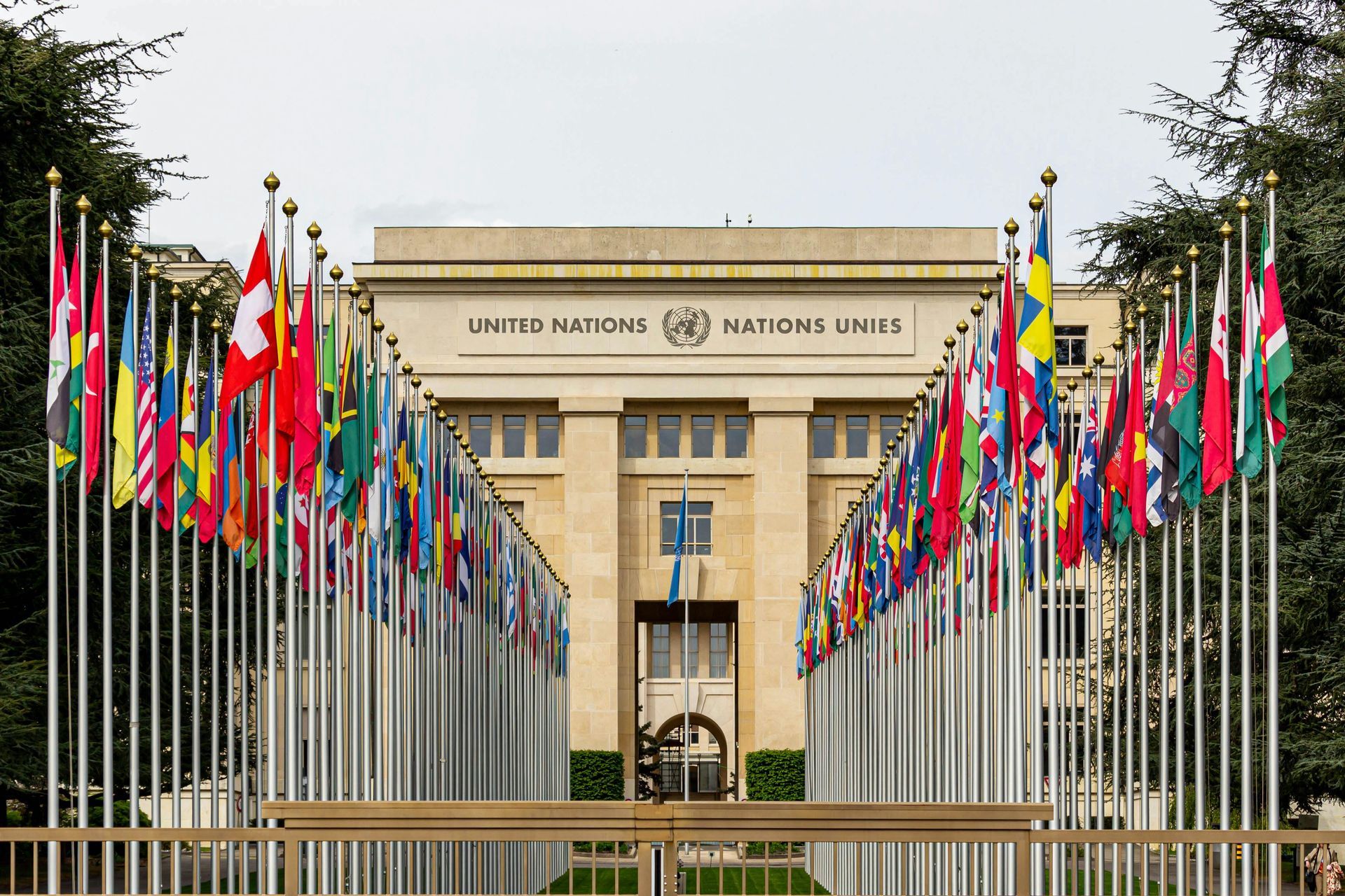 The United Nations Office at Geneva building behind two long rows of national flags flying on tall poles.