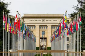 The United Nations Office at Geneva building behind two long rows of national flags flying on tall poles.