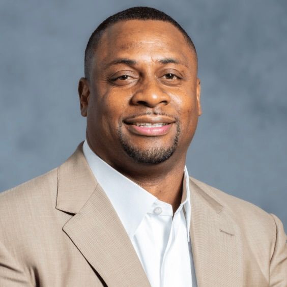 Headshot of a smiling person wearing a tan suit jacket and white collared shirt against a gray background.