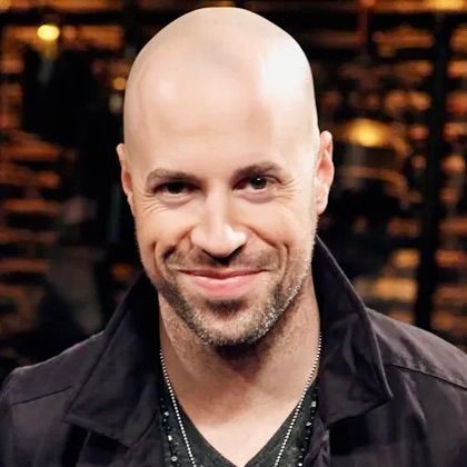 A bald man with a short beard smiles at the camera, wearing a black jacket and necklaces against a blurred background.