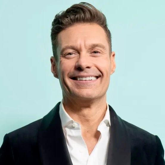 Ryan Seacrest smiling while wearing a dark blazer and white dress shirt against a light blue background.