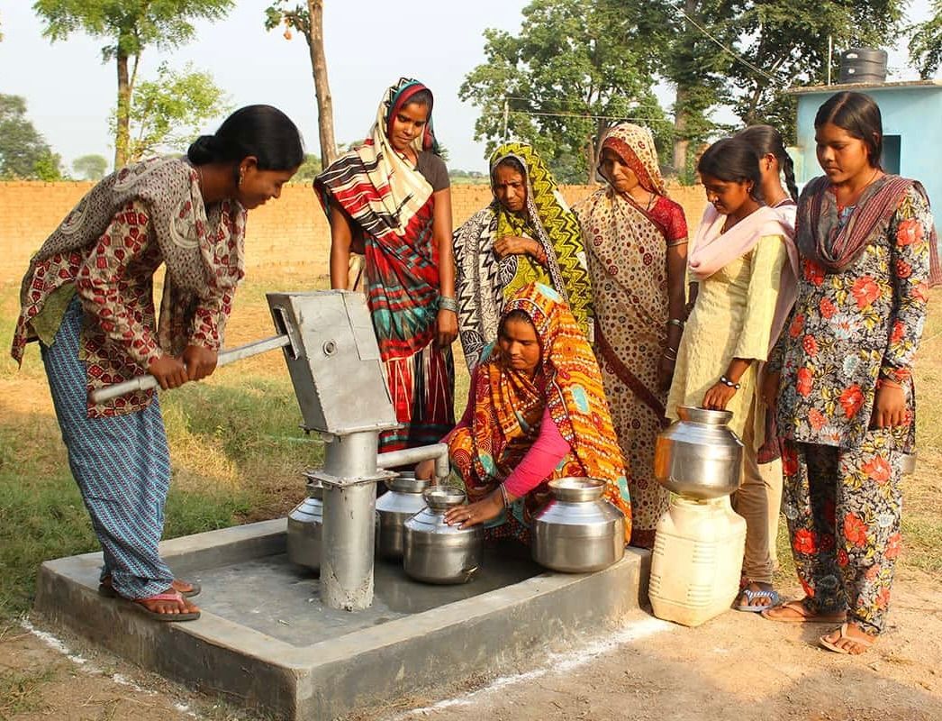 A group of people standing around a water pump, with one person pumping water into metal pots on a concrete base.