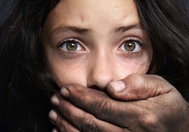 A close-up of a person with wide, expressive eyes having their mouth covered by a large, weathered hand.