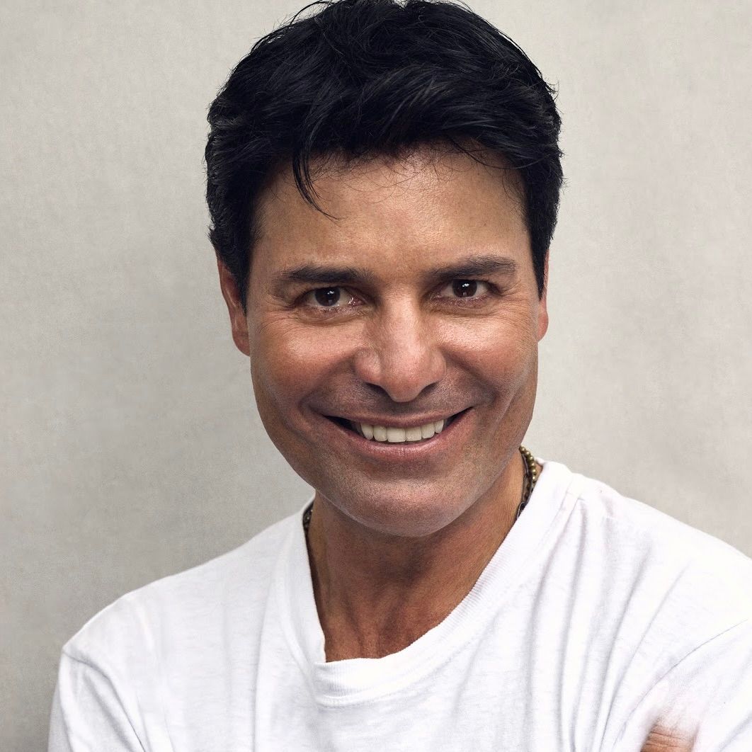 A headshot of a person with dark hair, smiling broadly while wearing a white shirt against a plain, light-colored background.