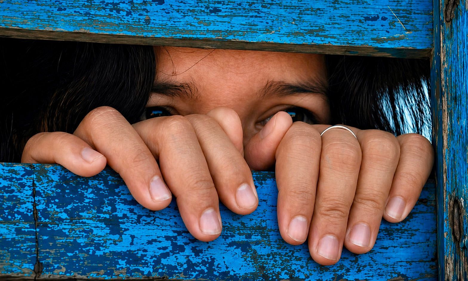A close-up of a person with wide, expressive eyes having their mouth covered by a large, weathered hand.