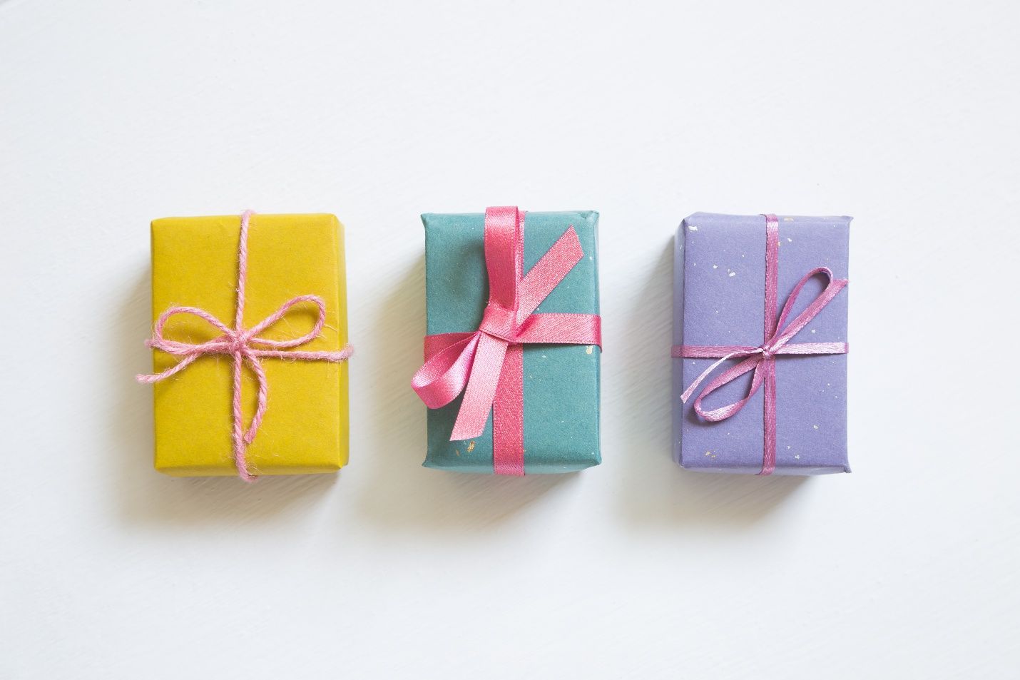Three wrapped gifts, yellow, teal, and purple, with pink ribbons, arranged on a white background.