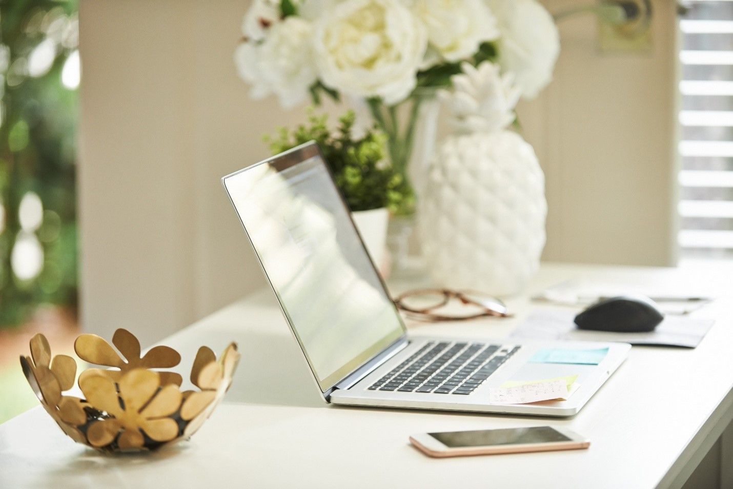 Laptop and phone on a white desk with flowers, a bowl, and pineapple vase.