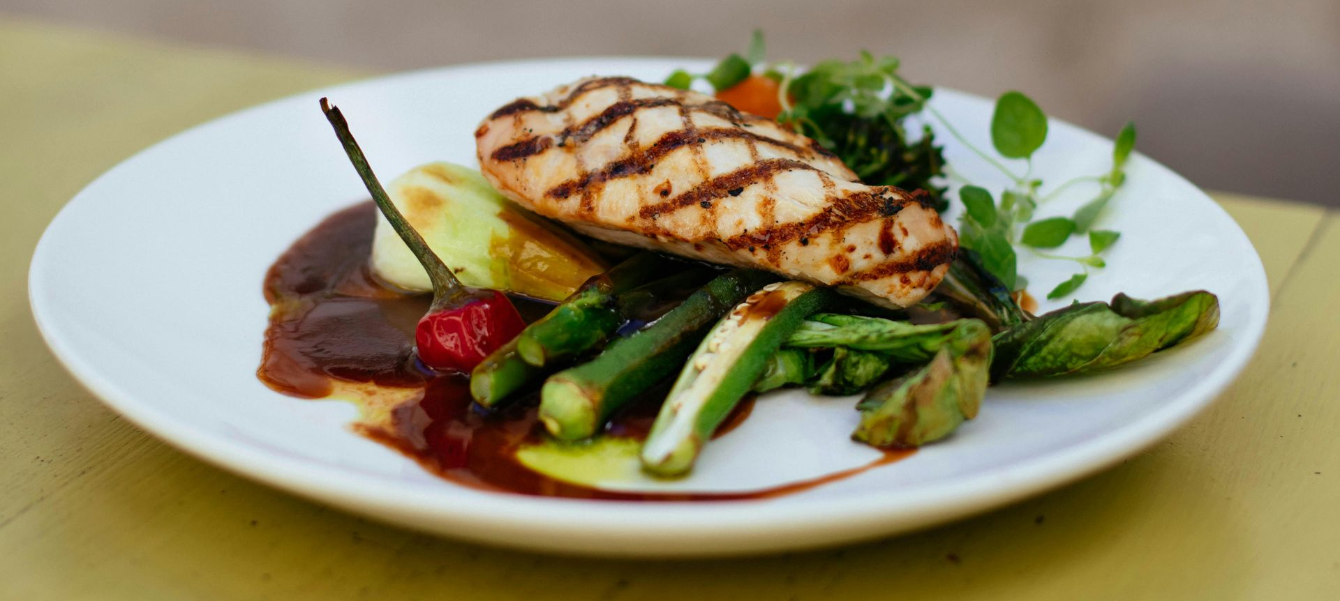 Grilled chicken breast with asparagus, okra, and sauce on a white plate.