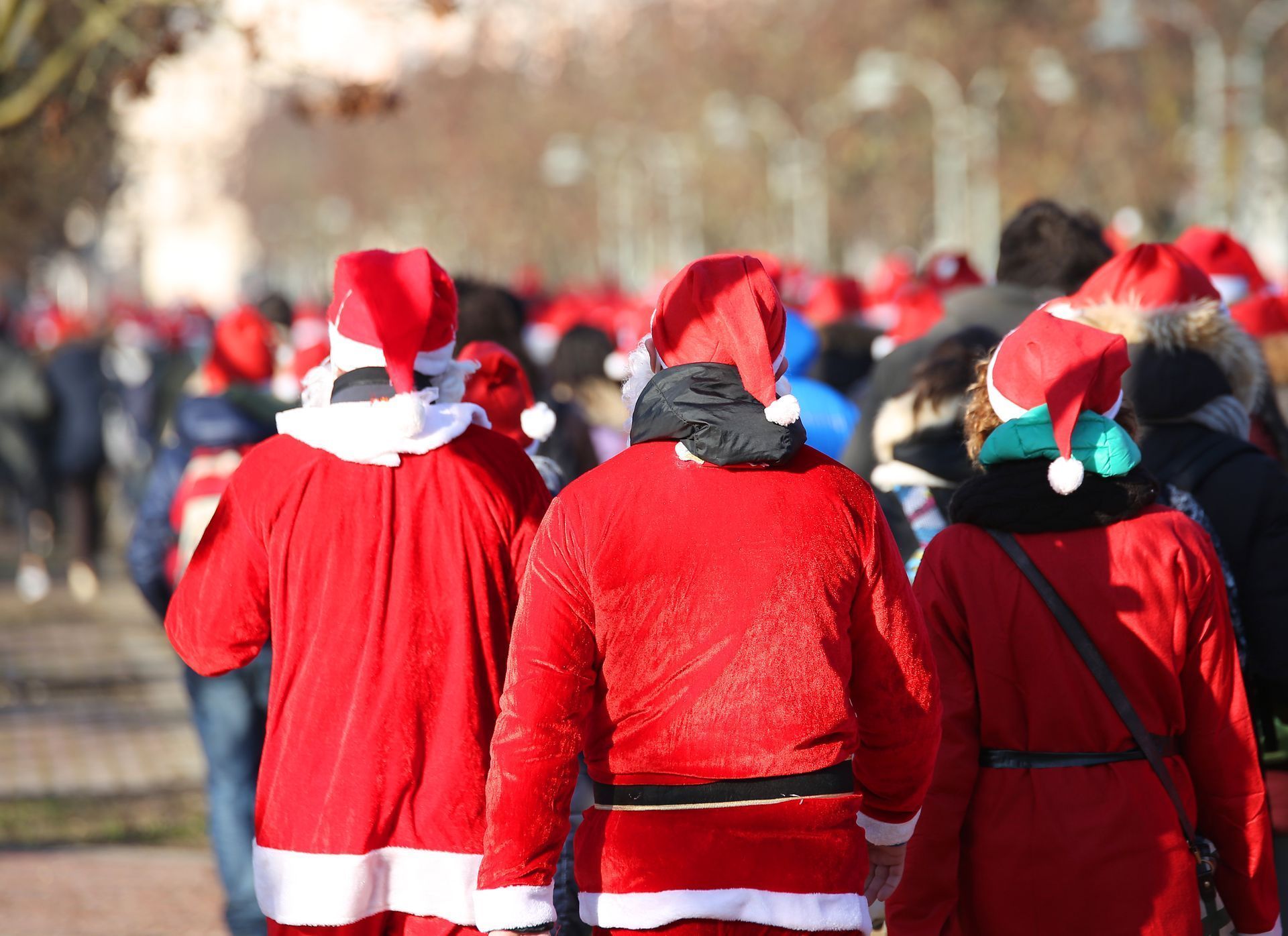 A large group of people dressed in Santa Claus costumes walk along a park pathway. The image is taken from behind, showing various people in festive red and white outfits.