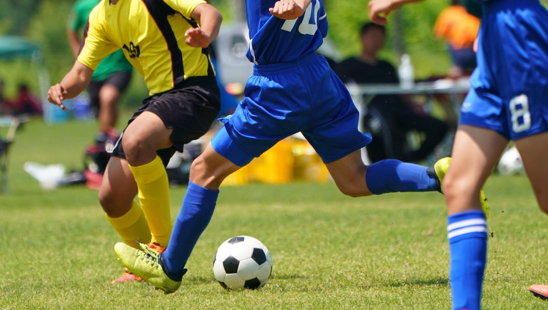 Players in yellow and blue jerseys chase a soccer ball on a grassy field during a daytime match. The focus is on the players' legs and the soccer ball.