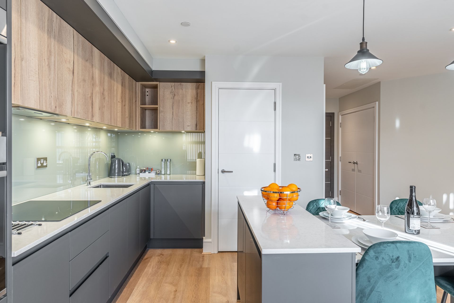 A bright kitchen with wood cabinets, white countertops. Sunlight streams in through large windows, illuminating the space.