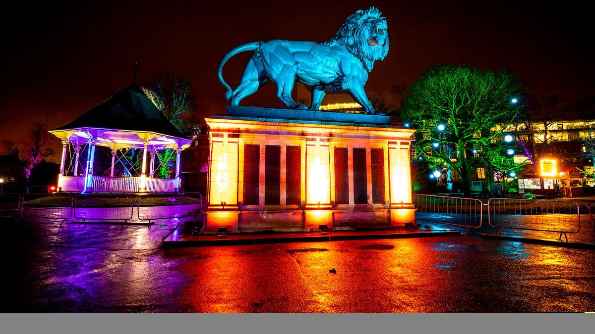 A large lion statue stands illuminated in vibrant colors at night next to a similarly lit pavilion, reflecting on the wet ground.