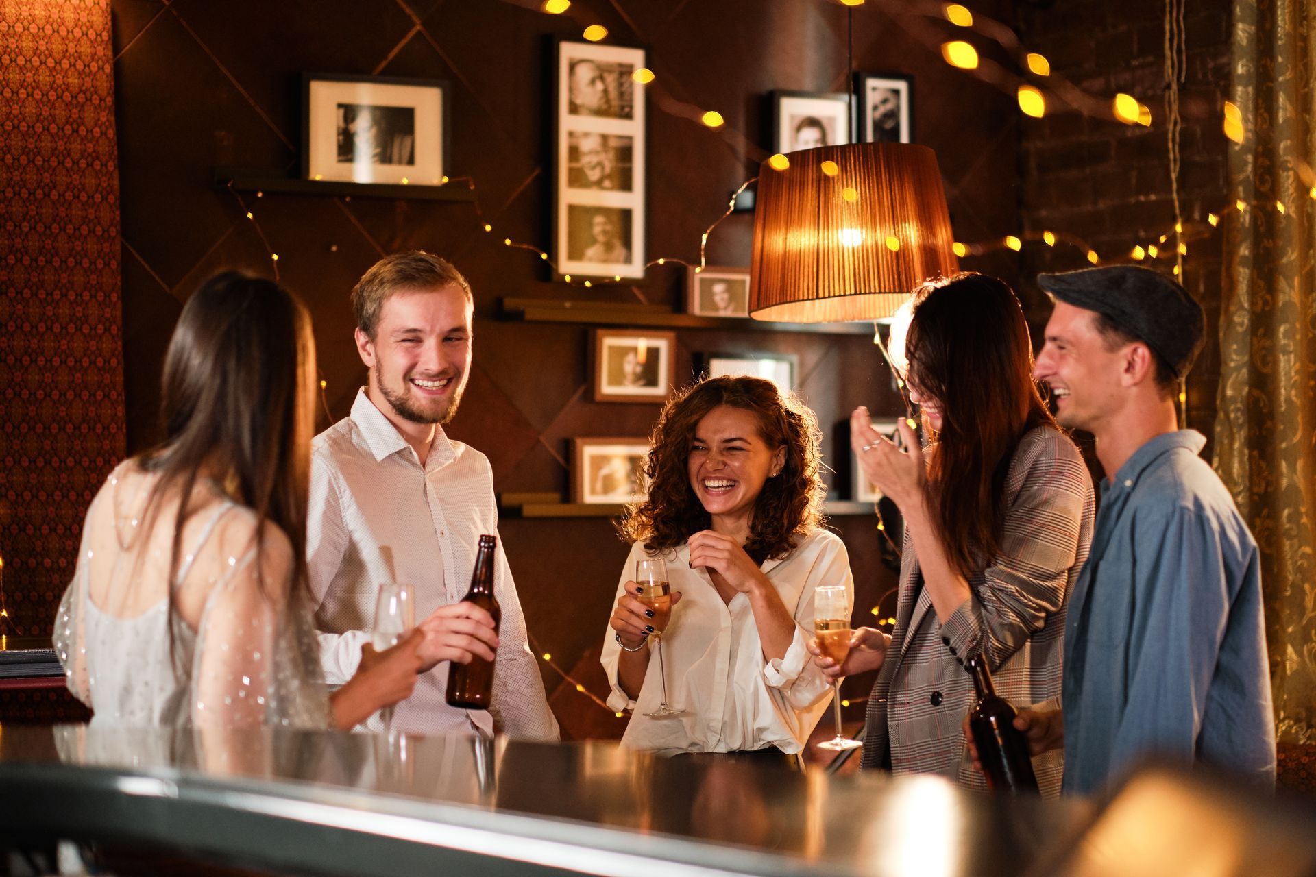 Five people socializing in a warmly lit room, holding drinks and smiling. Framed photos and string lights decorate the wall behind them.