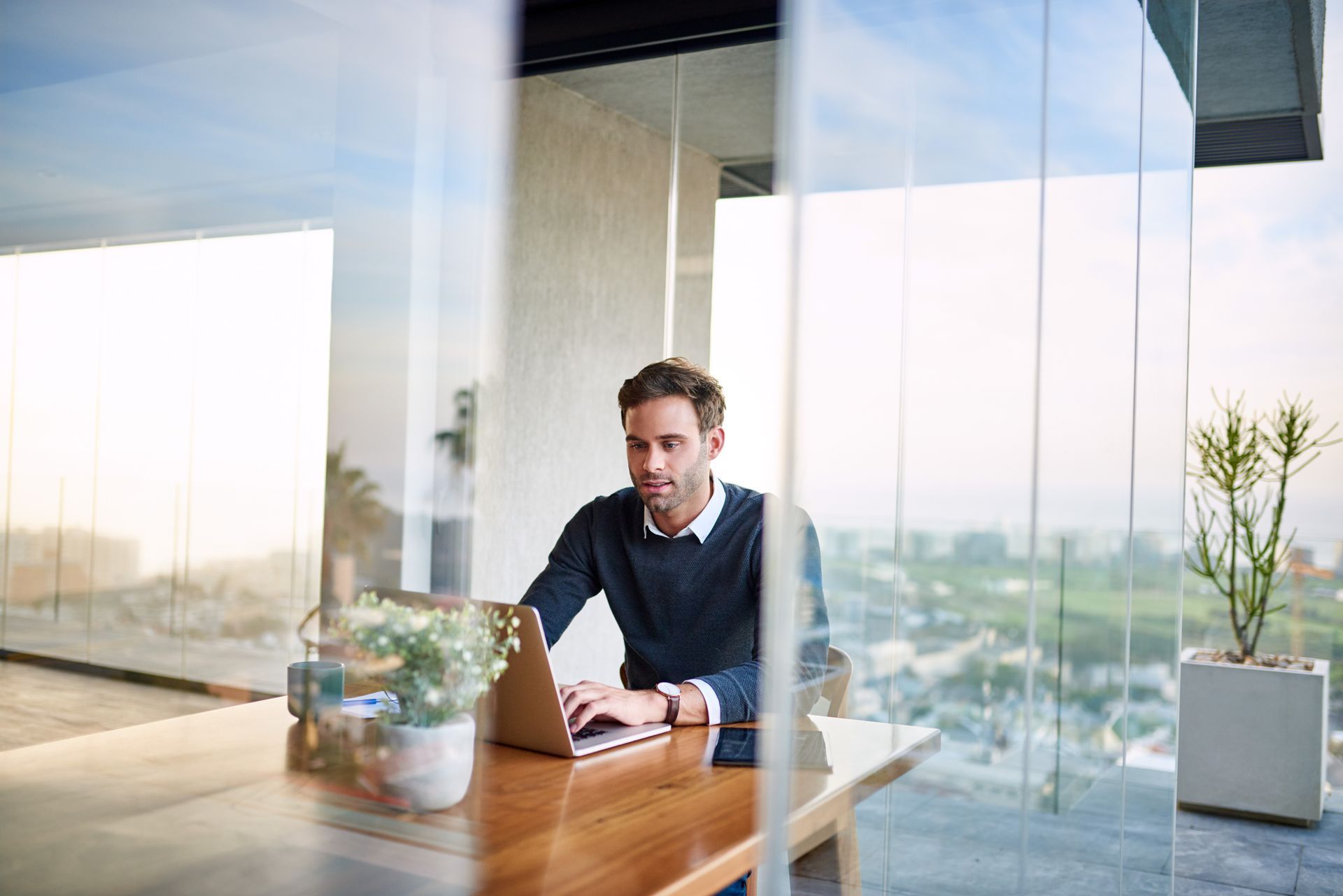 Man working on a laptop at a wooden table in a modern office with large windows and outdoor views.