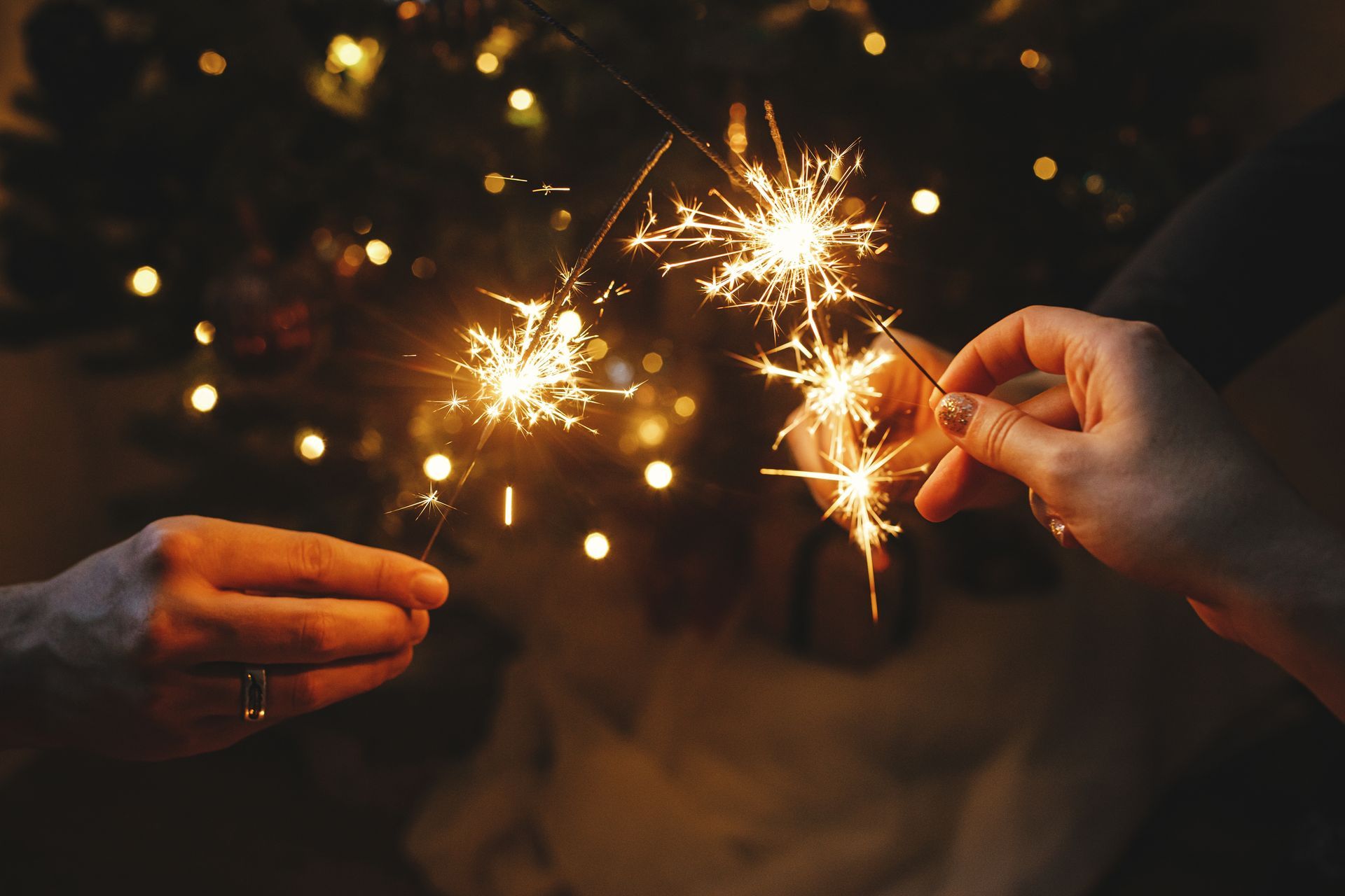 Three hands holding lit sparklers in front of a decorated Christmas tree with glowing lights capture the festive spirit, reminiscent of bonfire night in Reading.