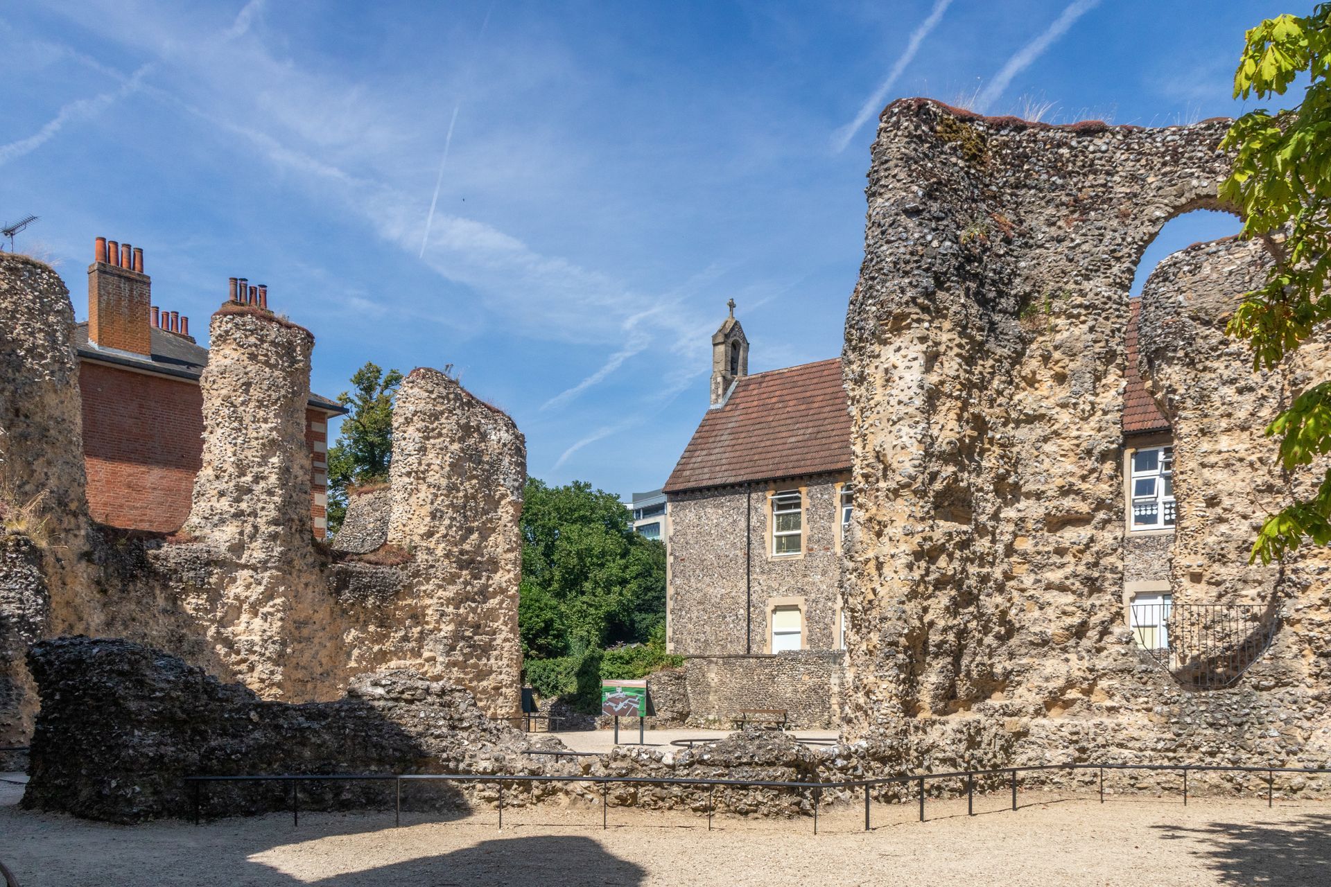 Ancient stone ruins with arched openings under a blue sky, surrounded by modern buildings and greenery.