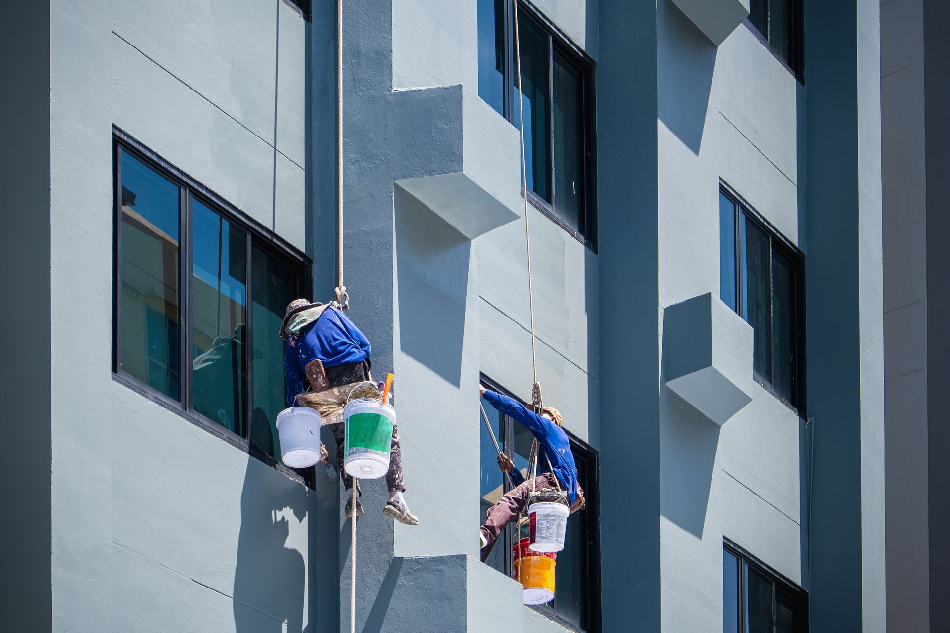 two men paint a building with buckets attached to ropes