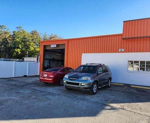 A red car and a blue SUV parked in front of an orange building with a white wall and a white fence.