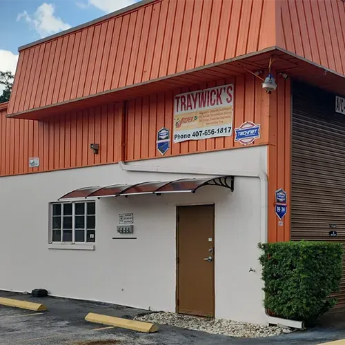 Exterior of Traywick's auto repair shop with orange roof and awning over a door, white walls, and a small bush.