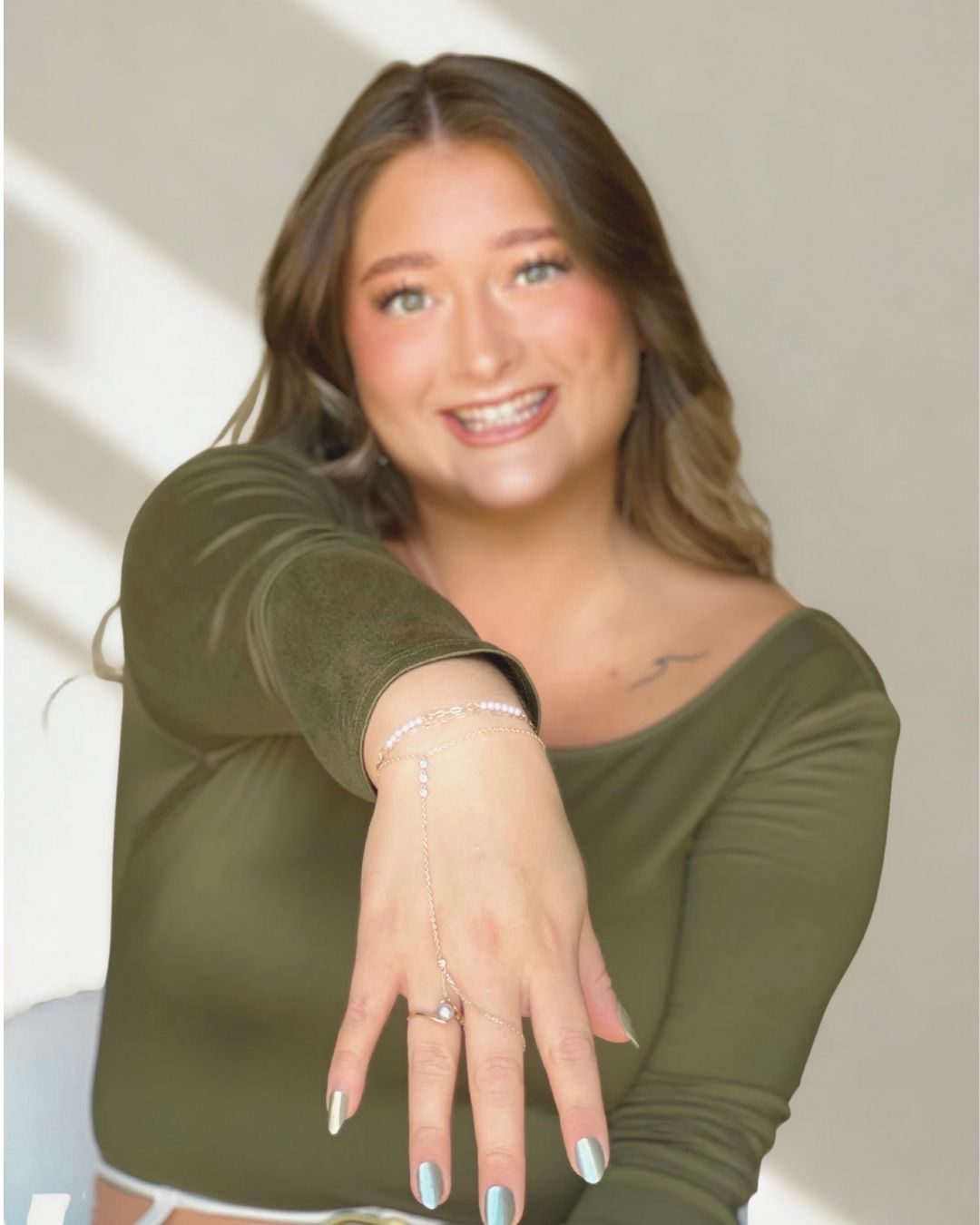 Woman in olive green top, holding hand up with ring, smiling.