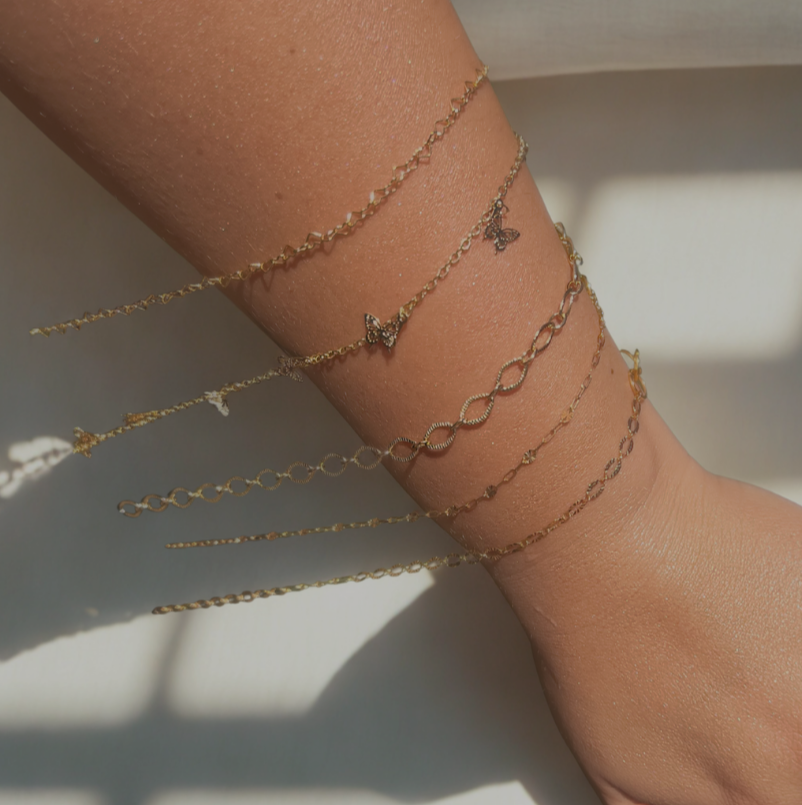 Gold bracelets on a wrist, various chains, some with charms, indoors, sunlight.
