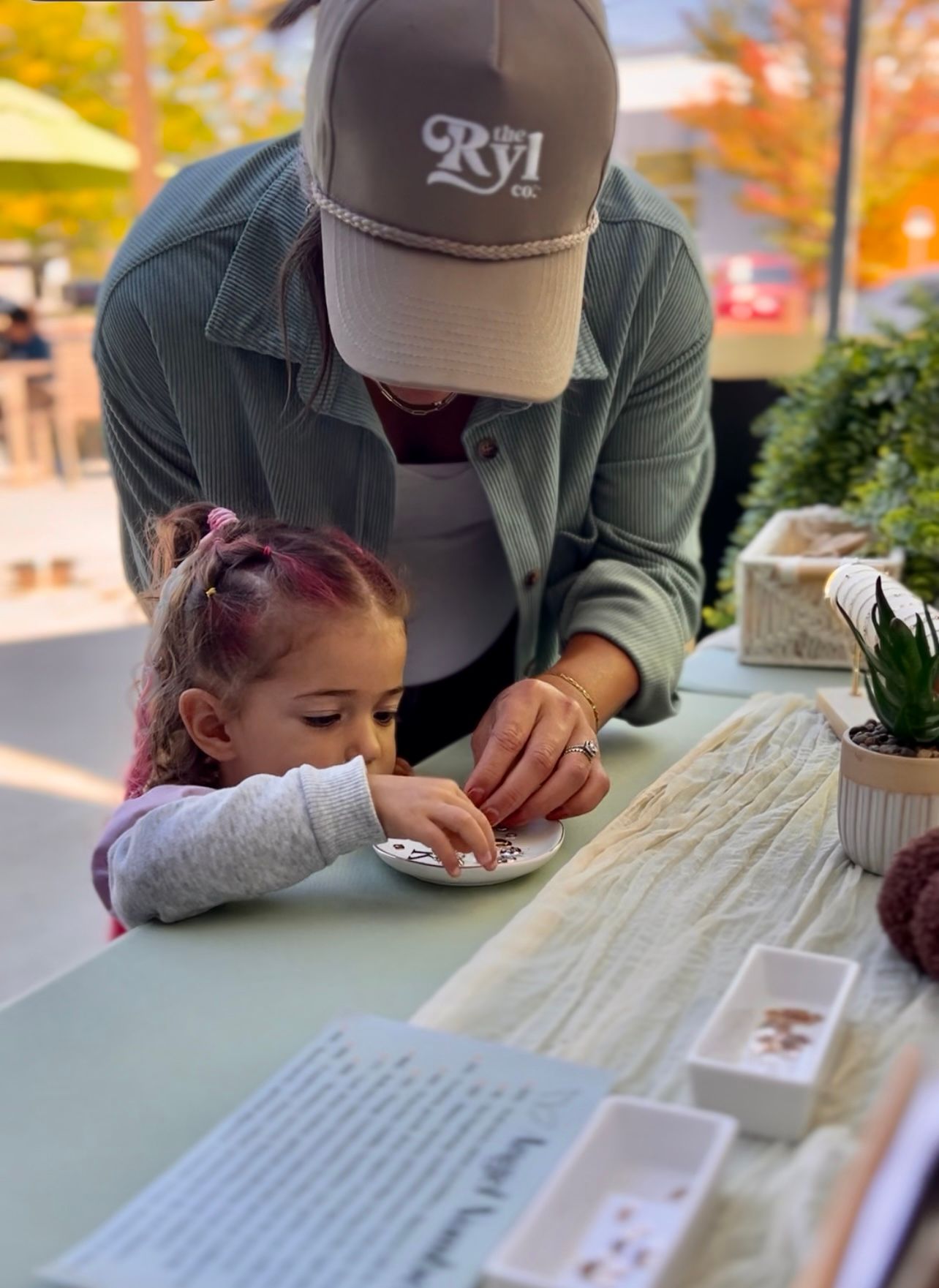 Woman and young girl looking at small objects on a table, outdoors. The girl is wearing a grey sweater.