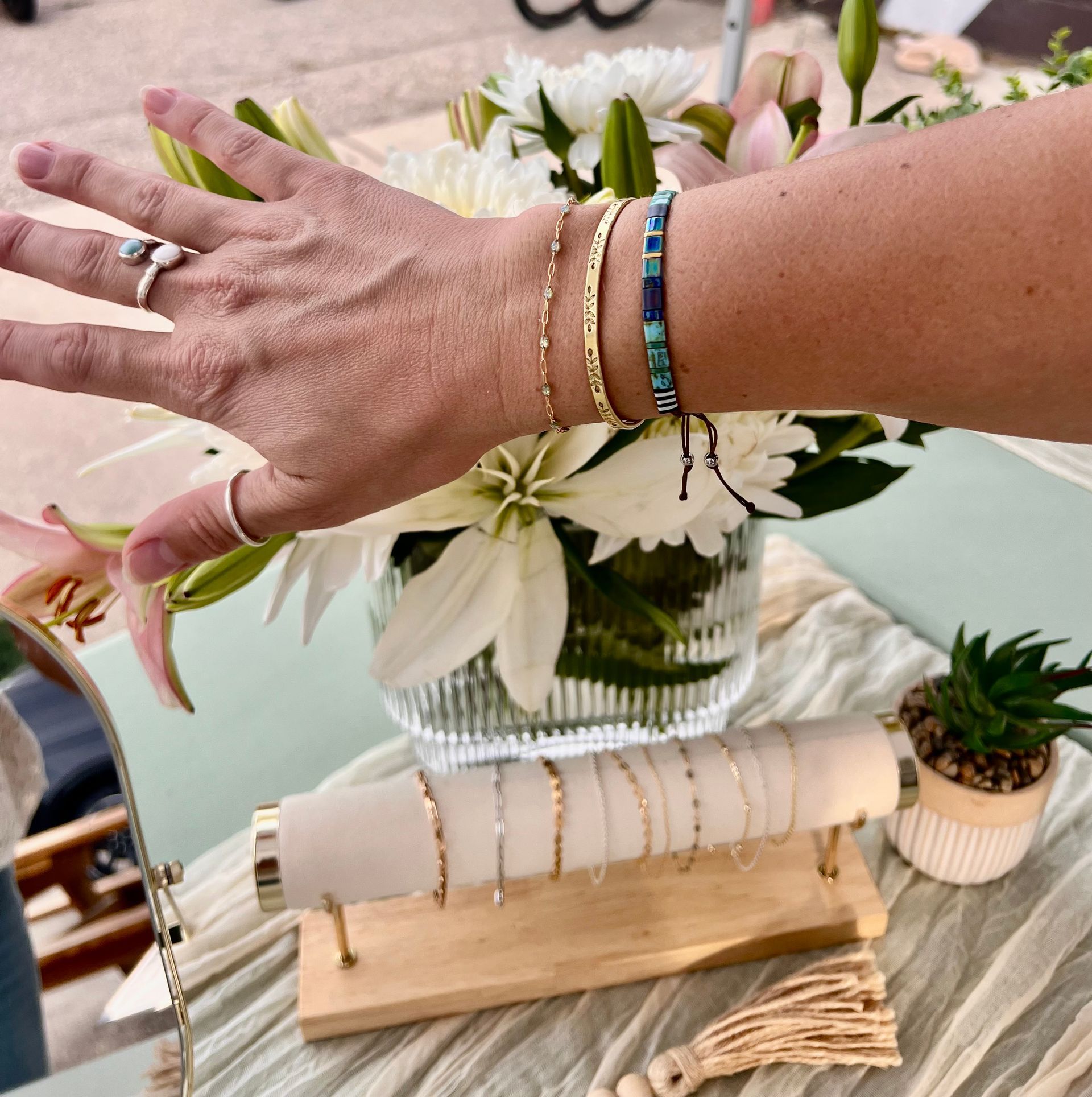 Woman's arm displaying gold bracelets, ring, and a turquoise bracelet near white flowers, and a display of more bracelets.
