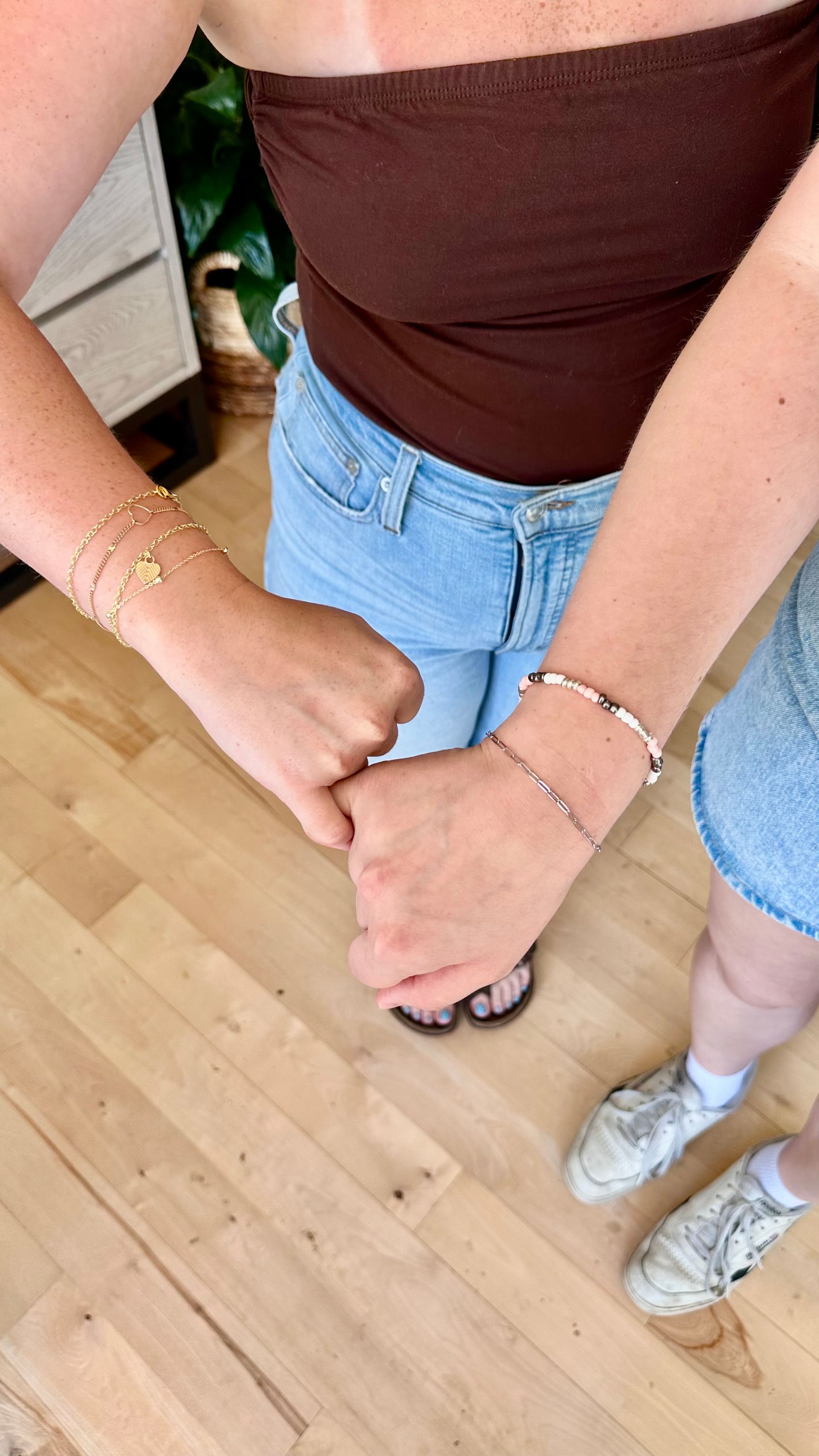 Two people holding hands, wearing bracelets, brown top and jean shorts, on wood floor.