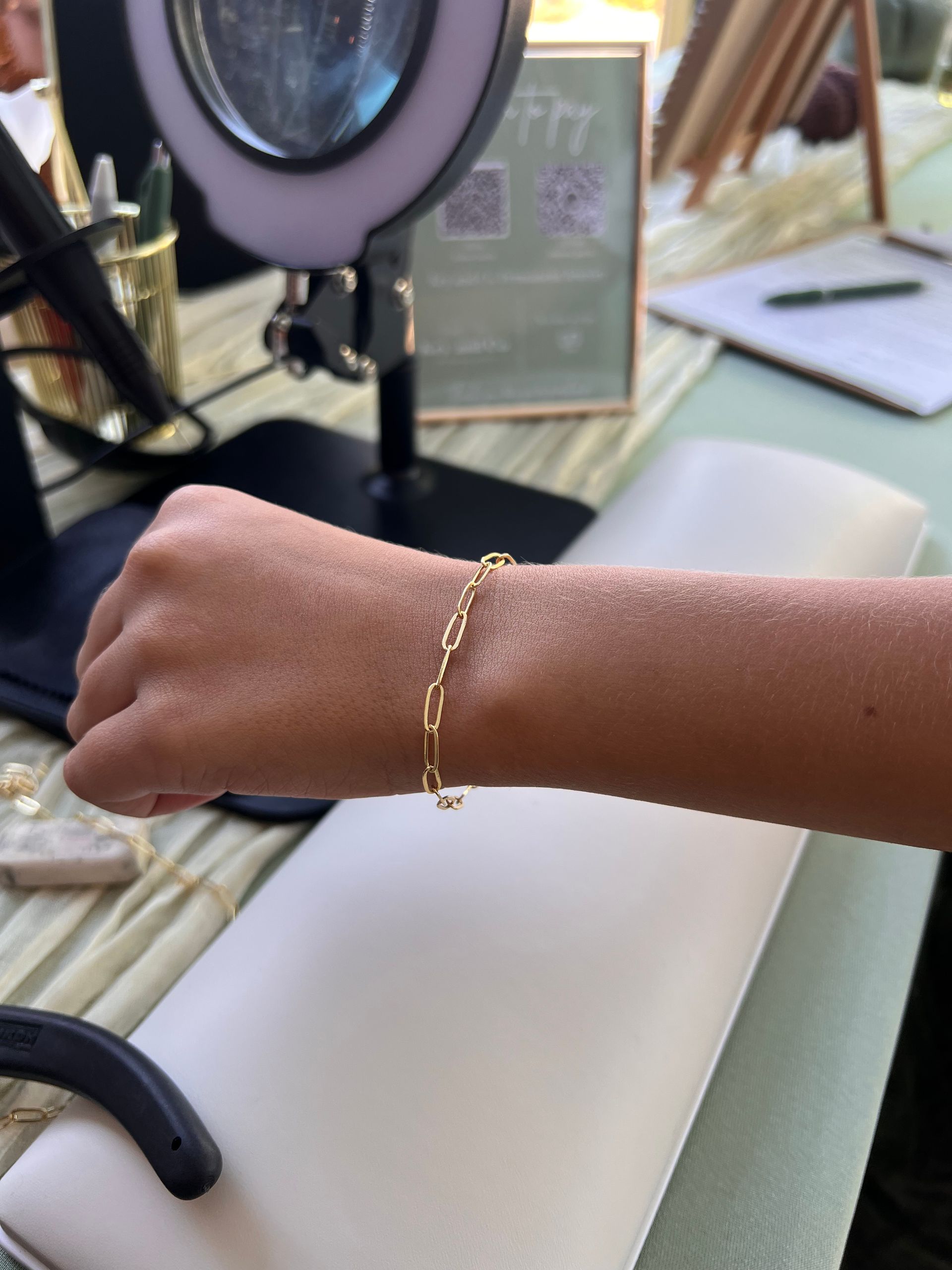 Person's arm with a gold chain bracelet, resting on a white surface. A ring light is in the background.
