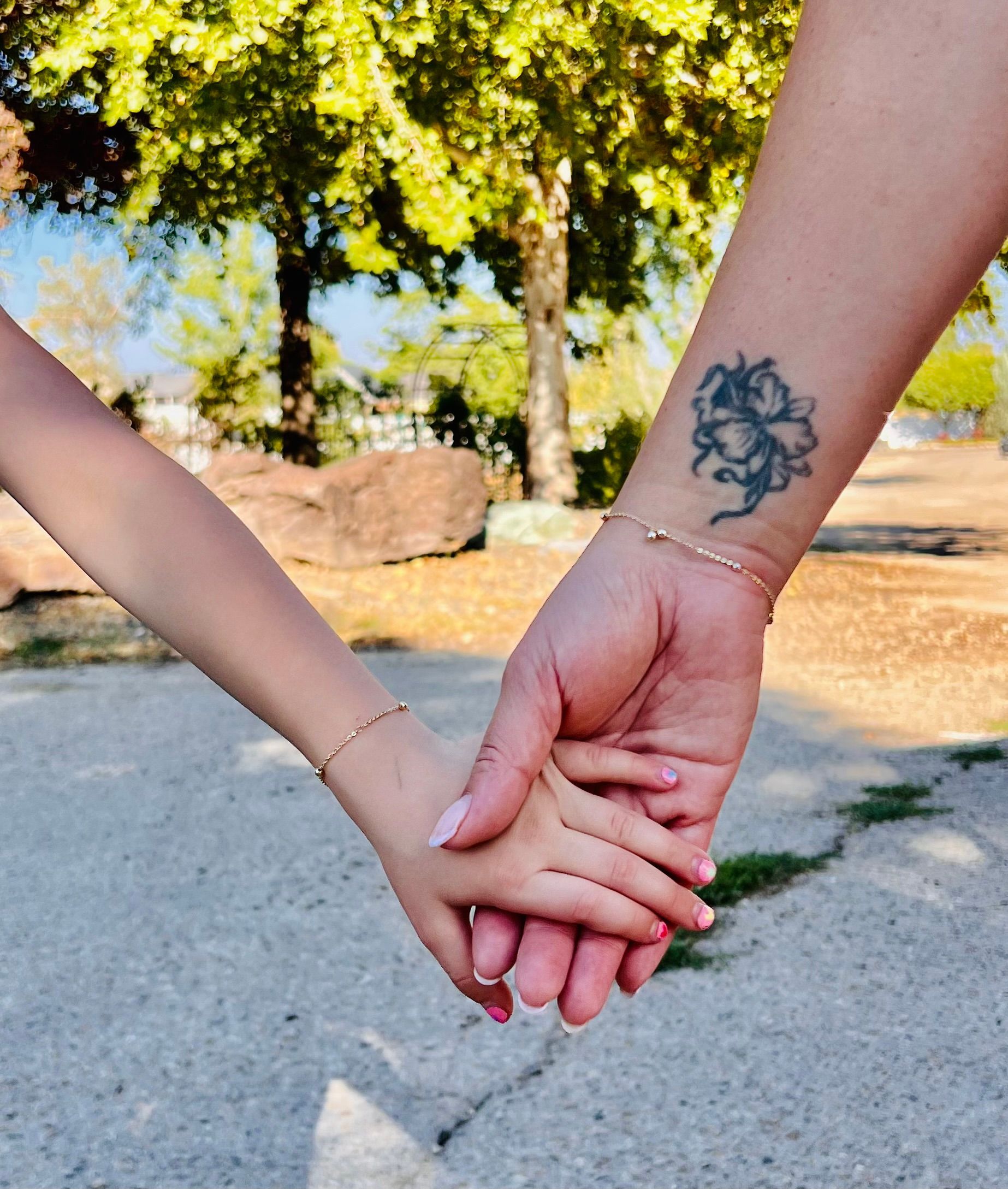 Adult and child hold hands in a park; both wearing gold bracelets. Adult has a wrist tattoo.