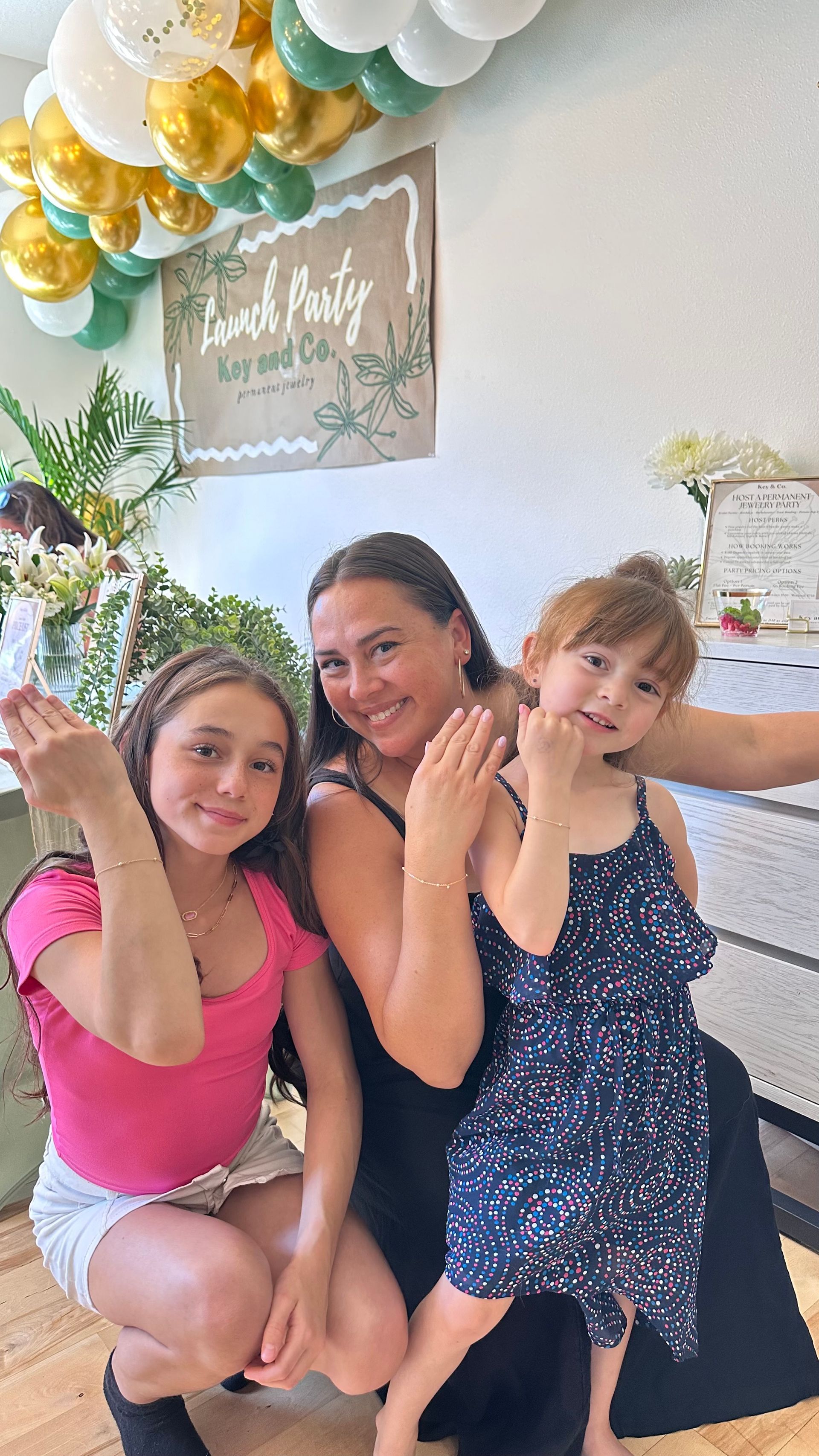 Woman and two young girls smiling, posing for a photo at a party with balloons and sign.