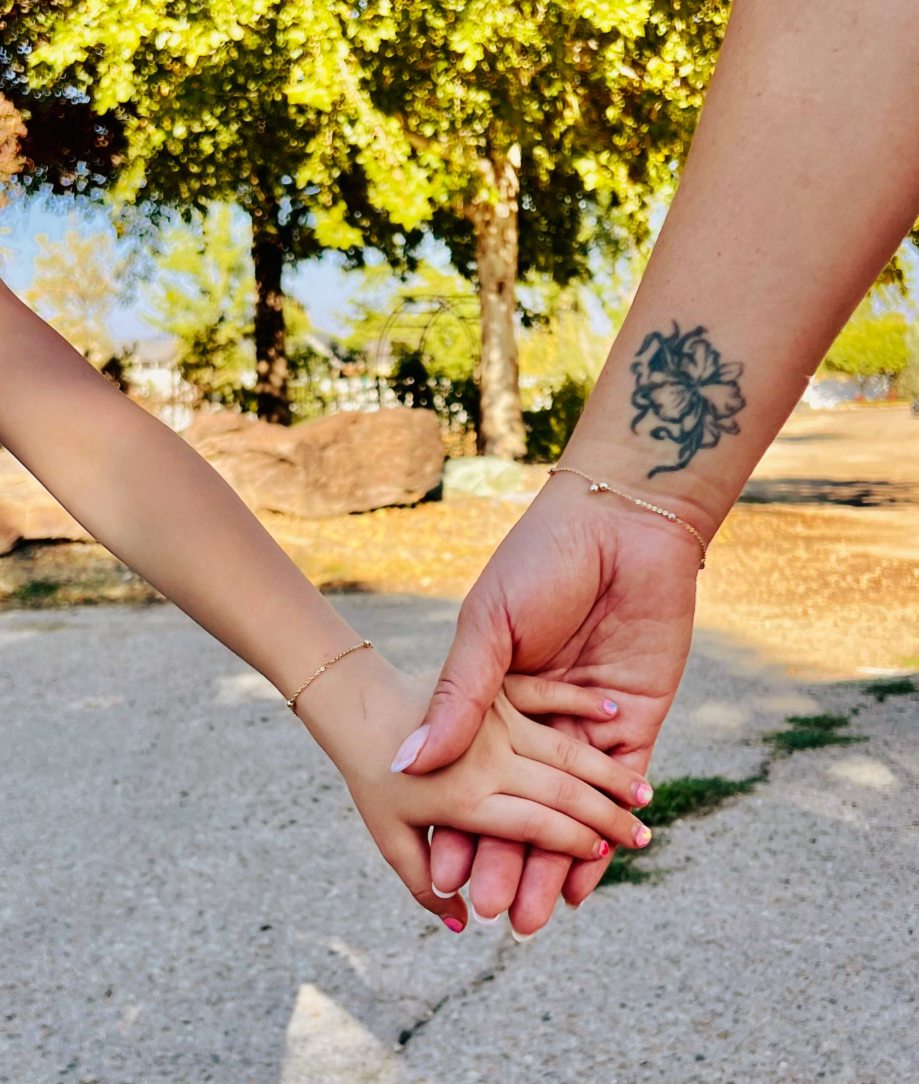 Adult and child hands clasped together outdoors; matching gold bracelets visible.