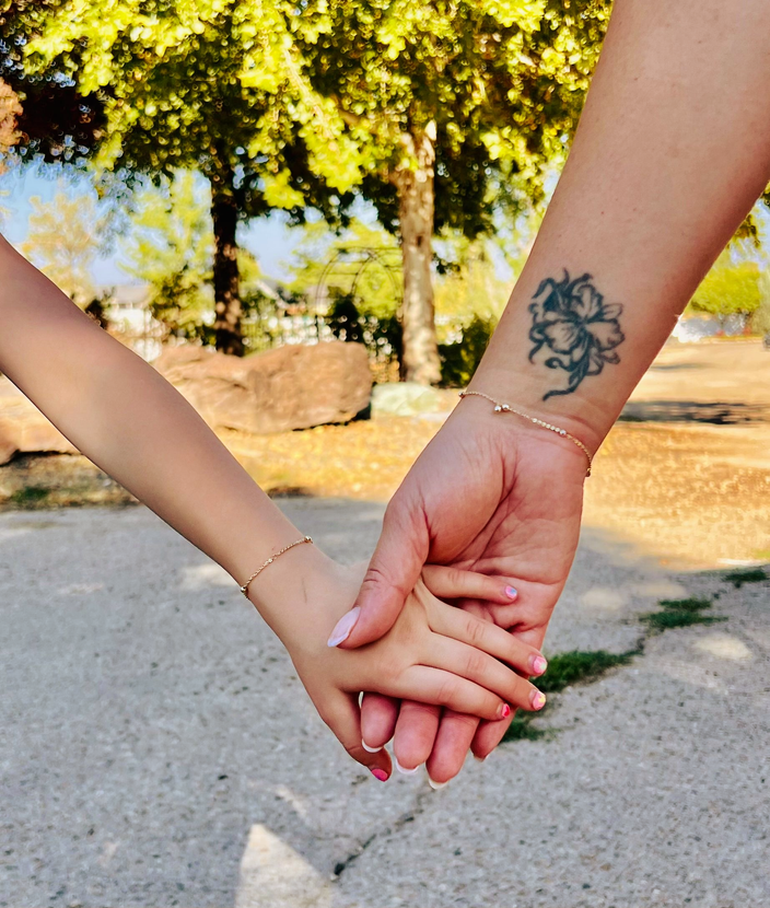 Adult and child hands clasped together outdoors; matching gold bracelets visible.