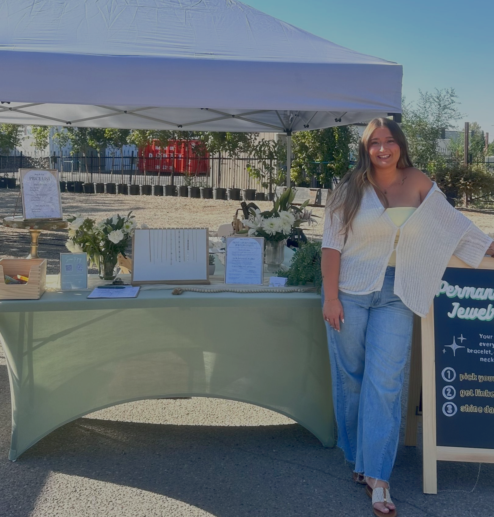 Woman at jewelry stand. She's wearing jeans, white top, smiling. Outdoor setting.