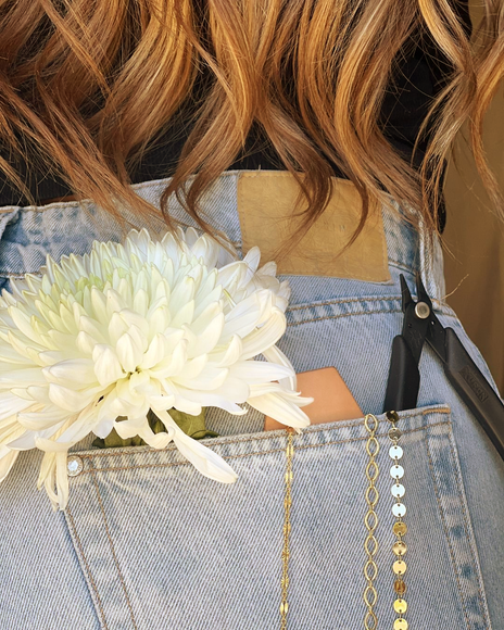 White flower, jewelry chain, and tools in denim back pocket; brown wavy hair in background.