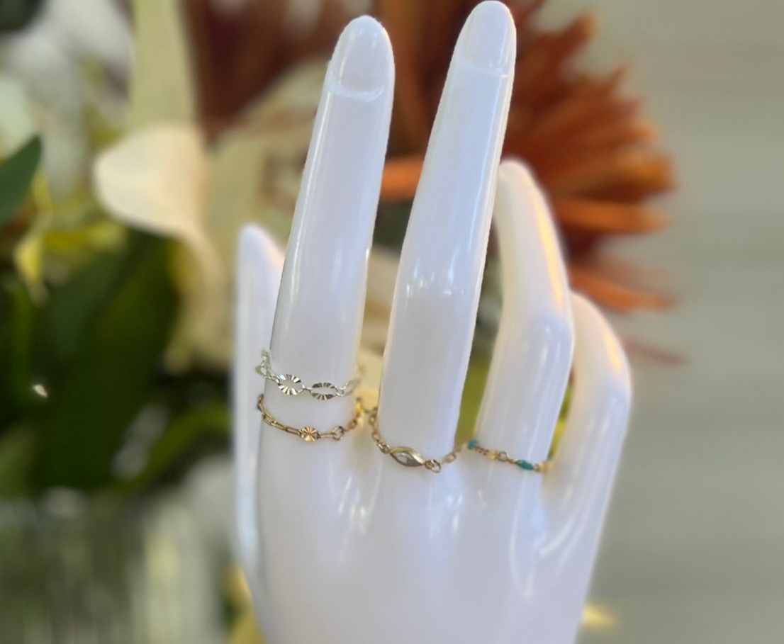 White hand mannequin with gold rings displayed in front of flowers.