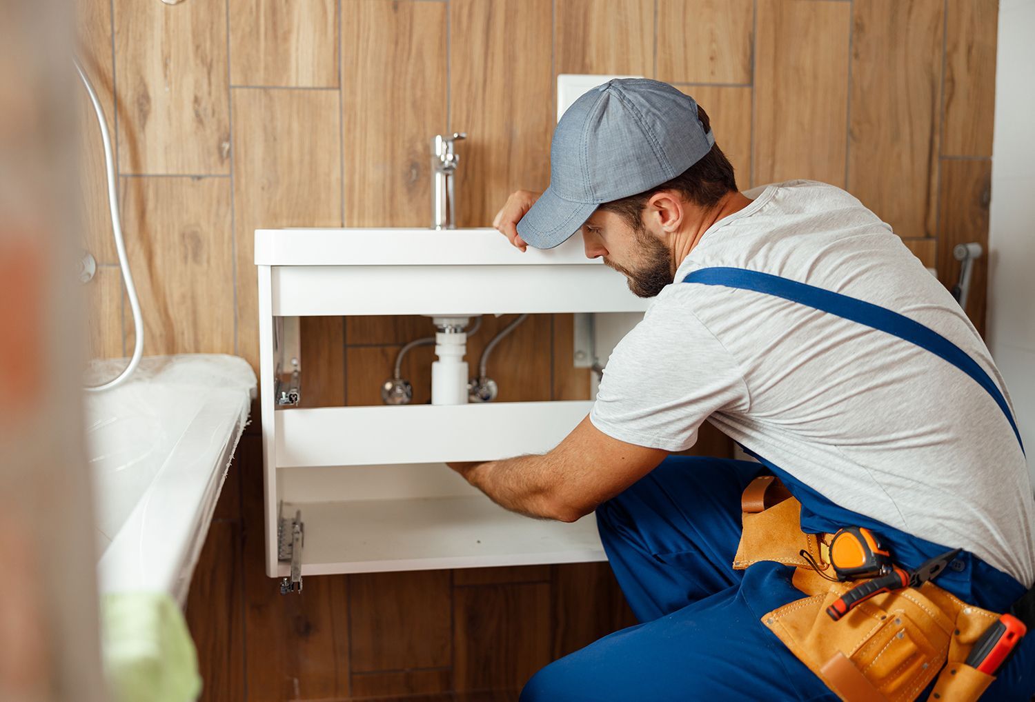 A male plumber installs a new pipe under a white sink, in a residential bathroom.